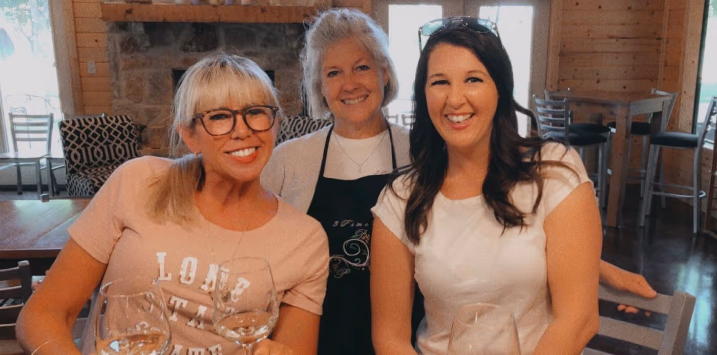 Three smiling women sitting at a wooden table with wine glasses, inside a cozy room with wooden walls and fireplace.