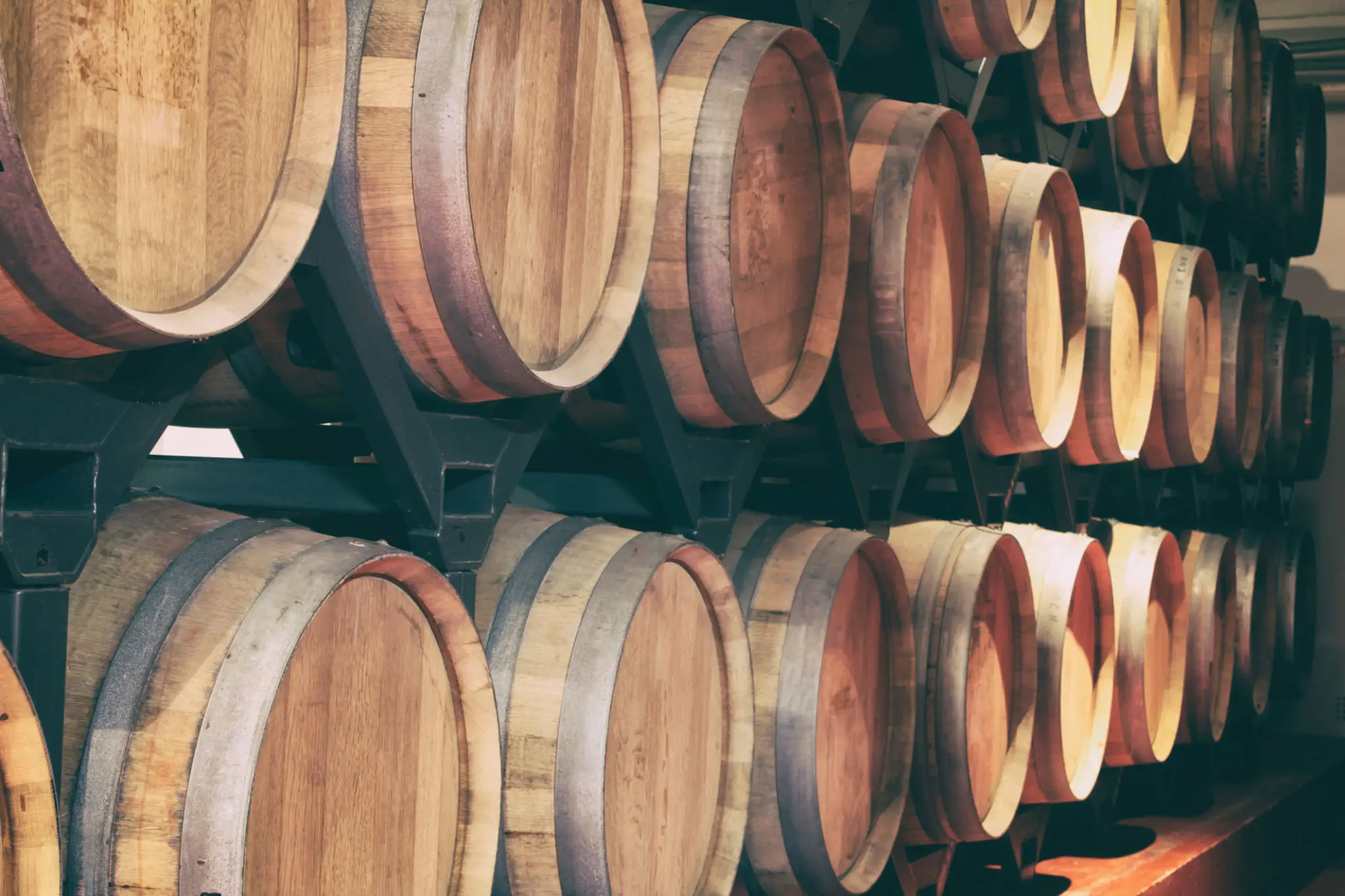 Rows of wooden wine barrels stacked on metal racks in a dimly lit cellar.